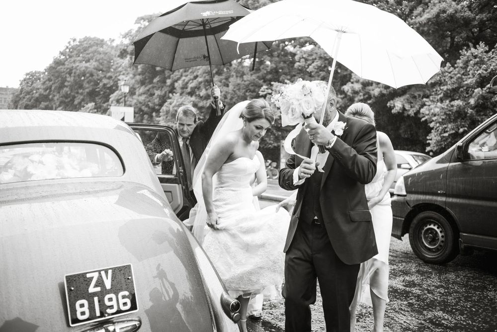 Bride and groom exit vintage car in rain holding umbrellas at outdoor wedding ceremony in black and white photo.