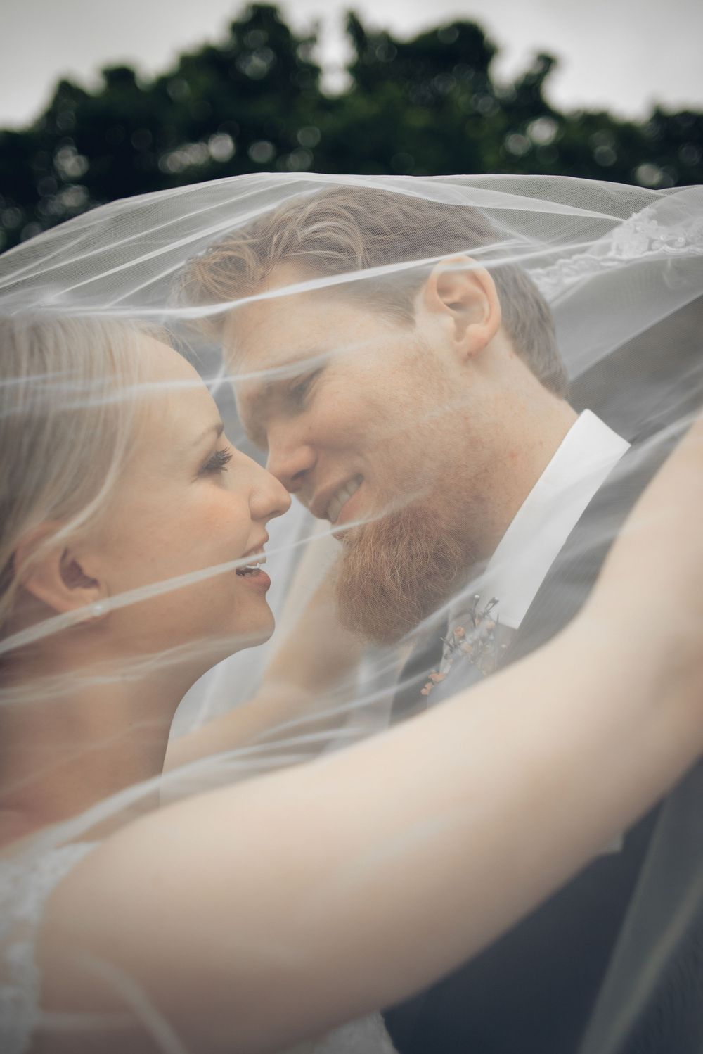 Bride and groom sharing a romantic moment under a wedding veil, smiling at each other outdoors.