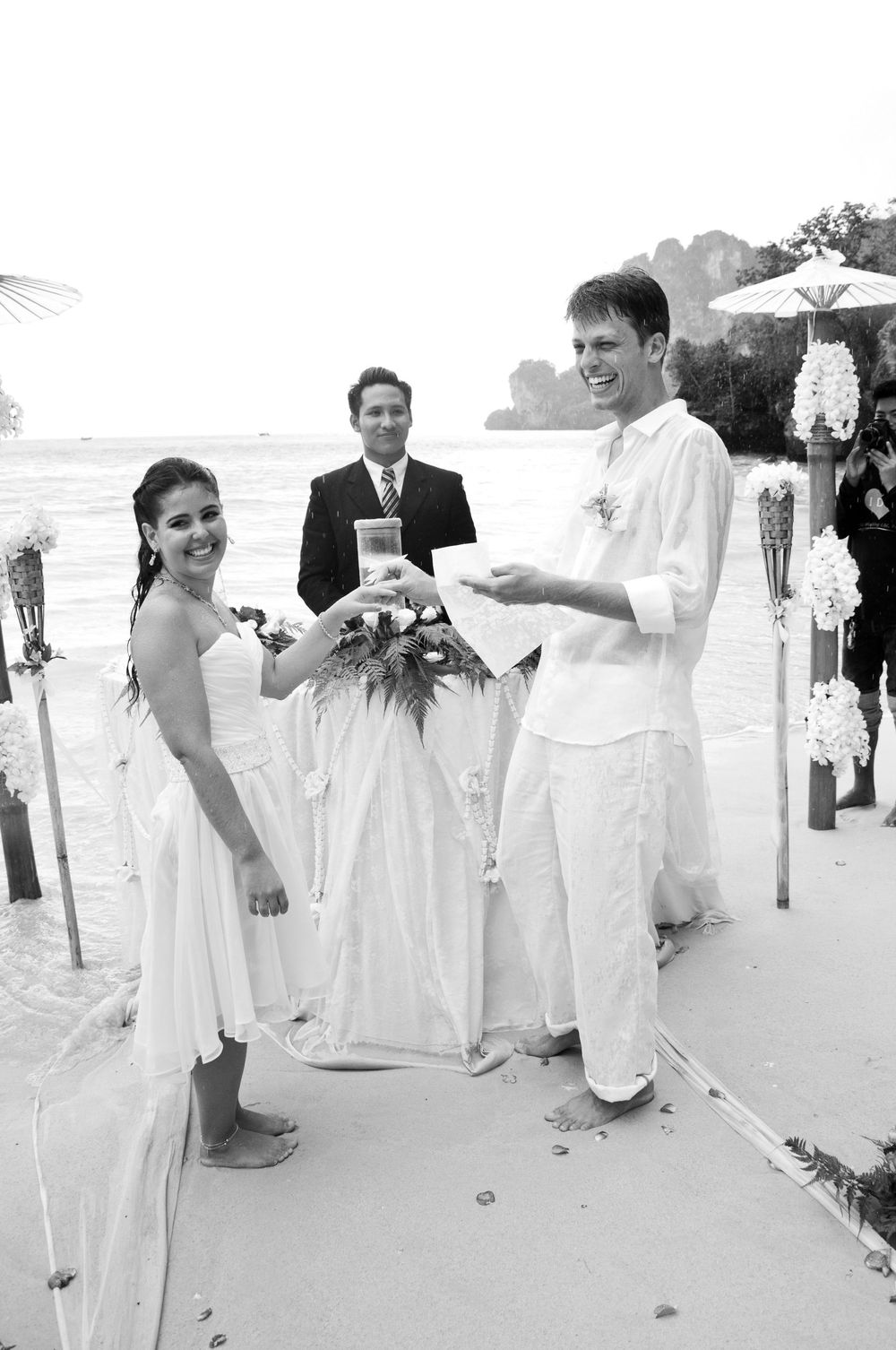 Black and white beach wedding ceremony with smiling bride and groom holding flowers, officiant standing behind them.