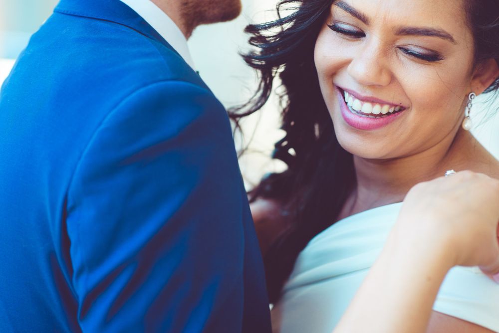 A radiant bride in a white dress smiles joyfully beside a groom in a blue suit at a wedding.