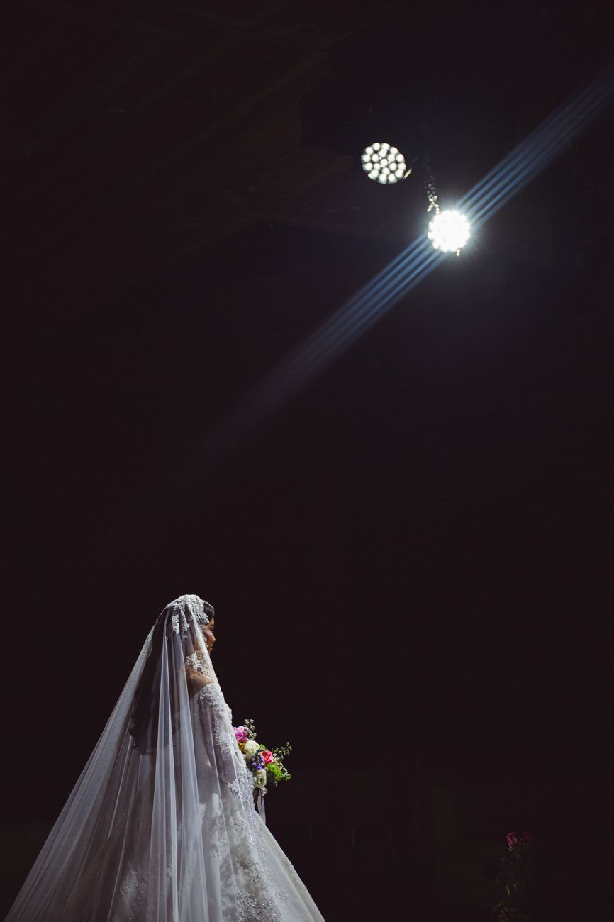 Bride in white lace gown and veil holds bouquet under dramatic spotlight against dark background.