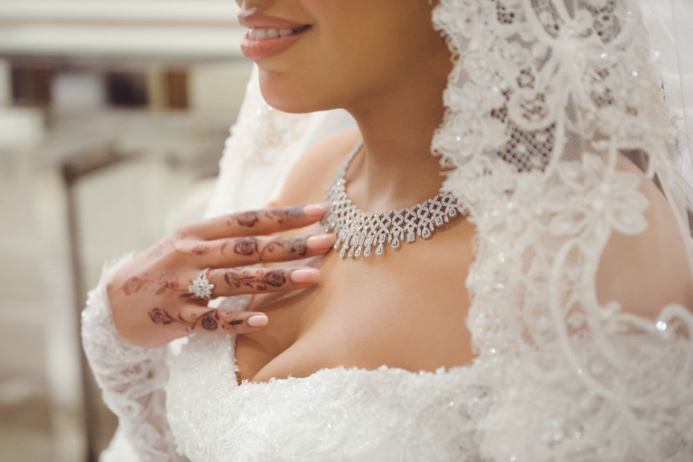 Bride in white lace gown with henna hands, diamond necklace, and veil on wedding day.