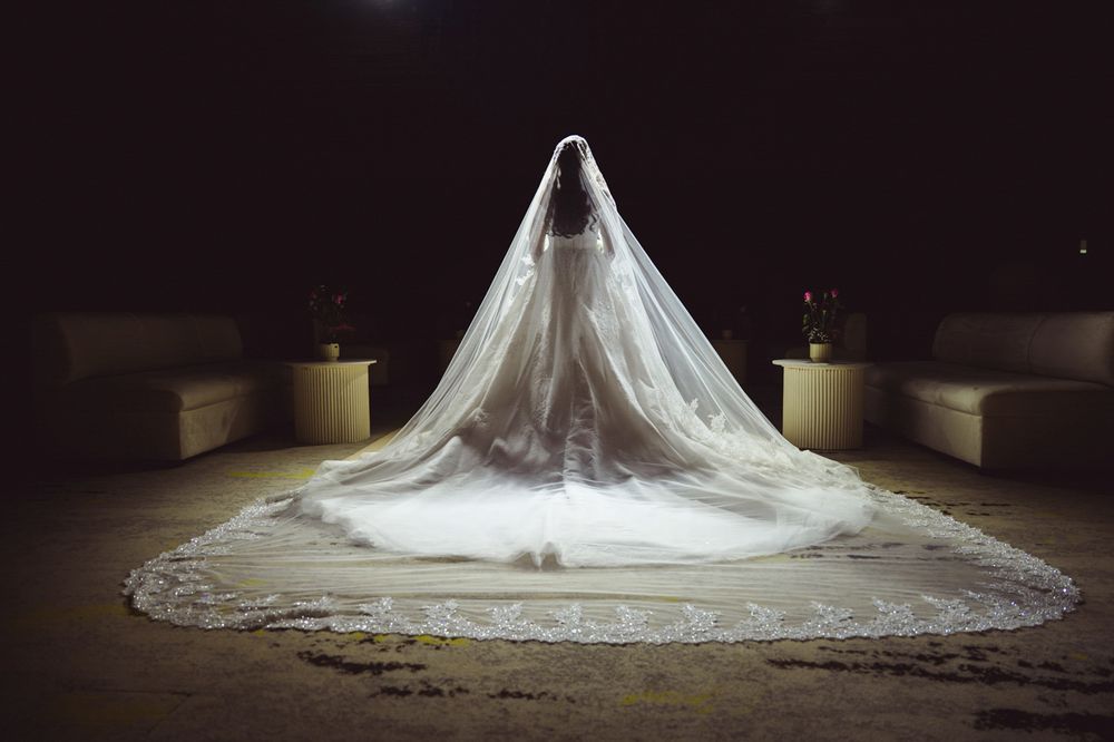 Bride in a stunning wedding gown with an elegant lace-trimmed cathedral veil spread dramatically on a dark floor.