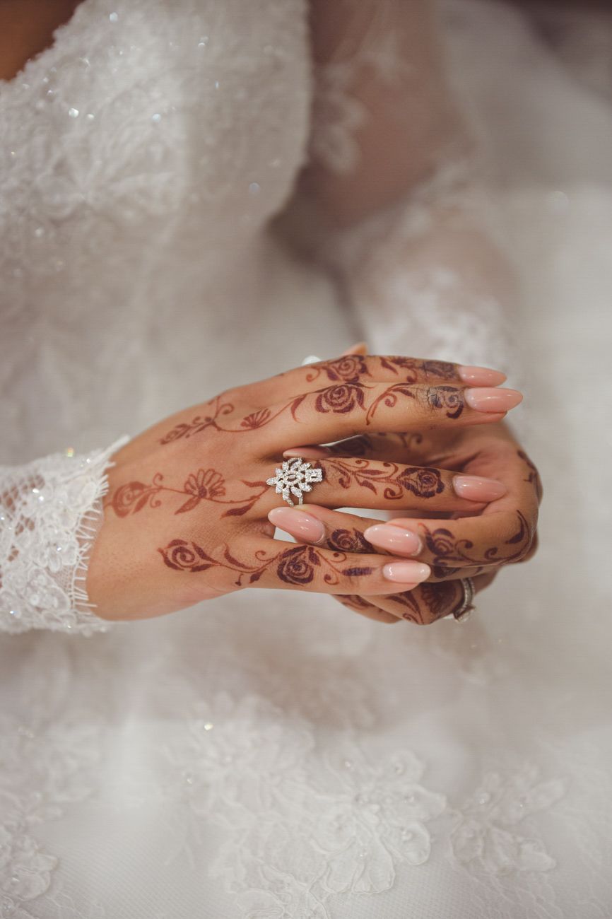 Bride's hands adorned with intricate henna tattoos and a diamond snowflake ring, wearing a white lace wedding dress.