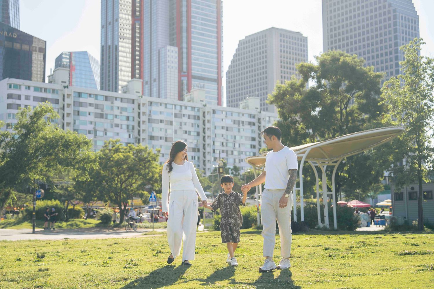 Family of three walking in urban park with city skyscrapers in background on a sunny day.