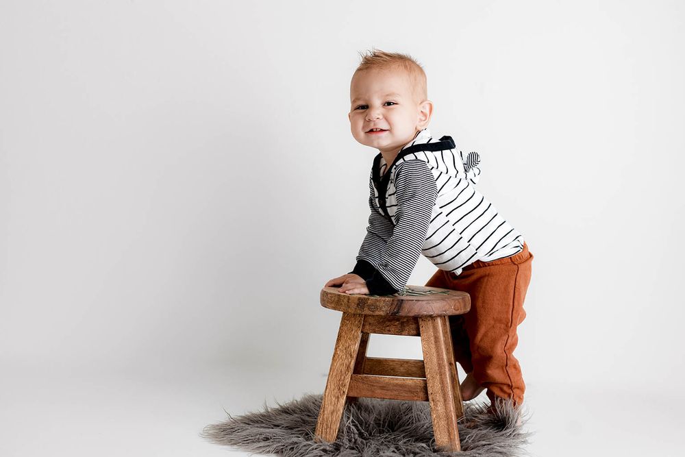 Smiling baby boy in striped shirt and rust pants climbing on rustic wooden stool on gray fur rug.