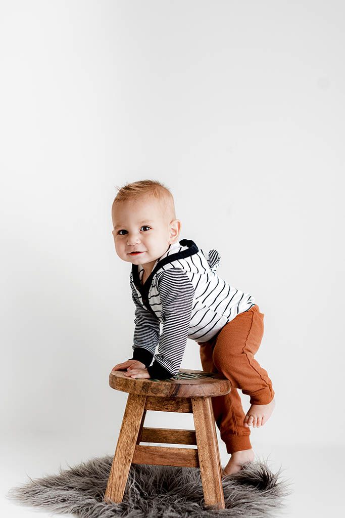 Adorable toddler in striped top and rust pants climbing wooden stool on fluffy gray rug, smiling.