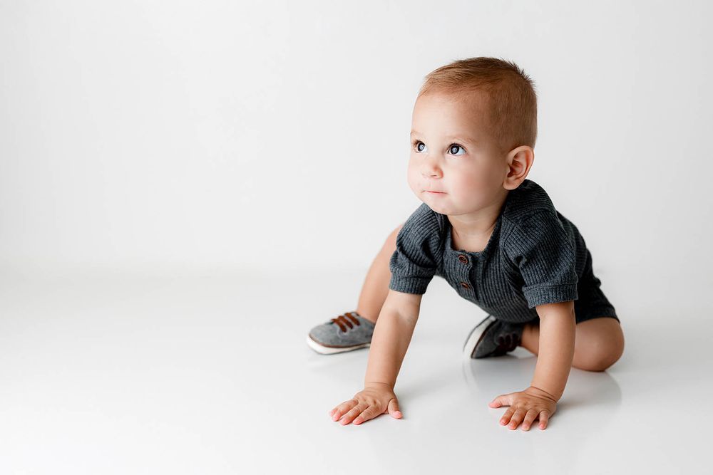 Red-haired baby boy crawling on white floor wearing gray onesie and sneakers.