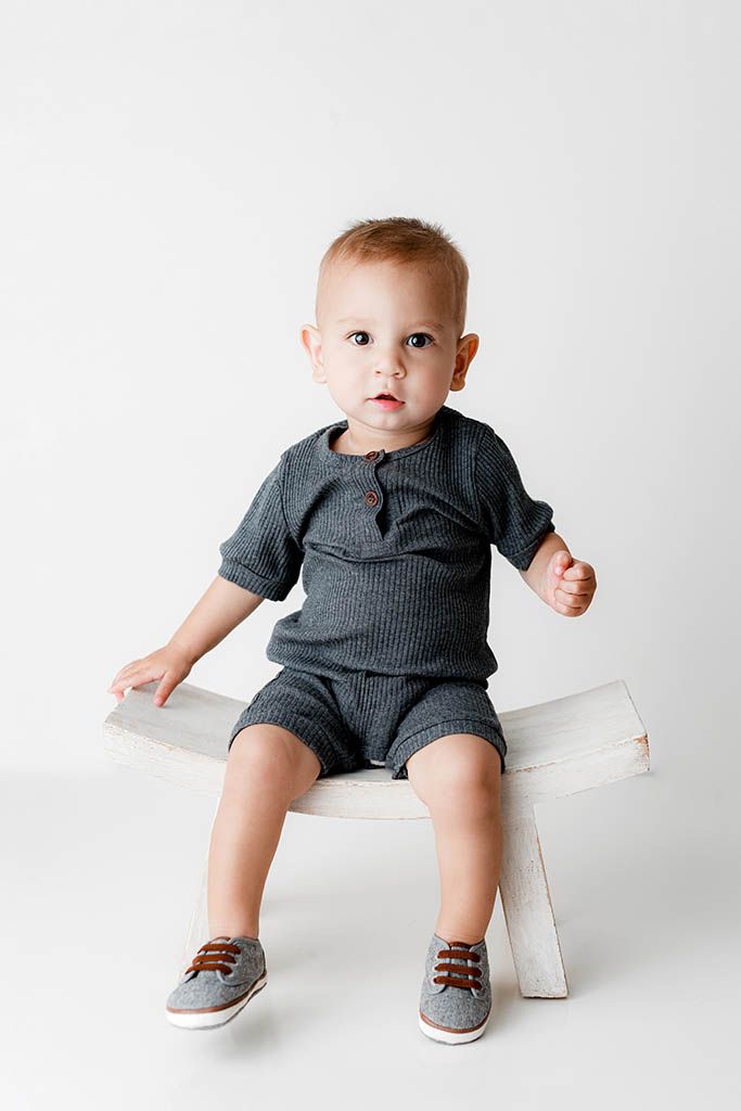Toddler boy sitting on white bench wearing gray ribbed matching shorts set and gray sneakers with brown laces.