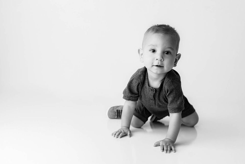 Black and white photo of a cute baby boy crawling on a white studio floor, looking at the camera.