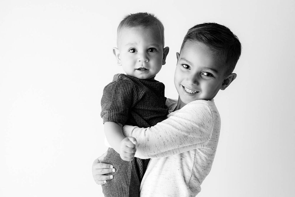 Black and white photo of an older boy smiling and holding a baby sibling in a studio portrait.