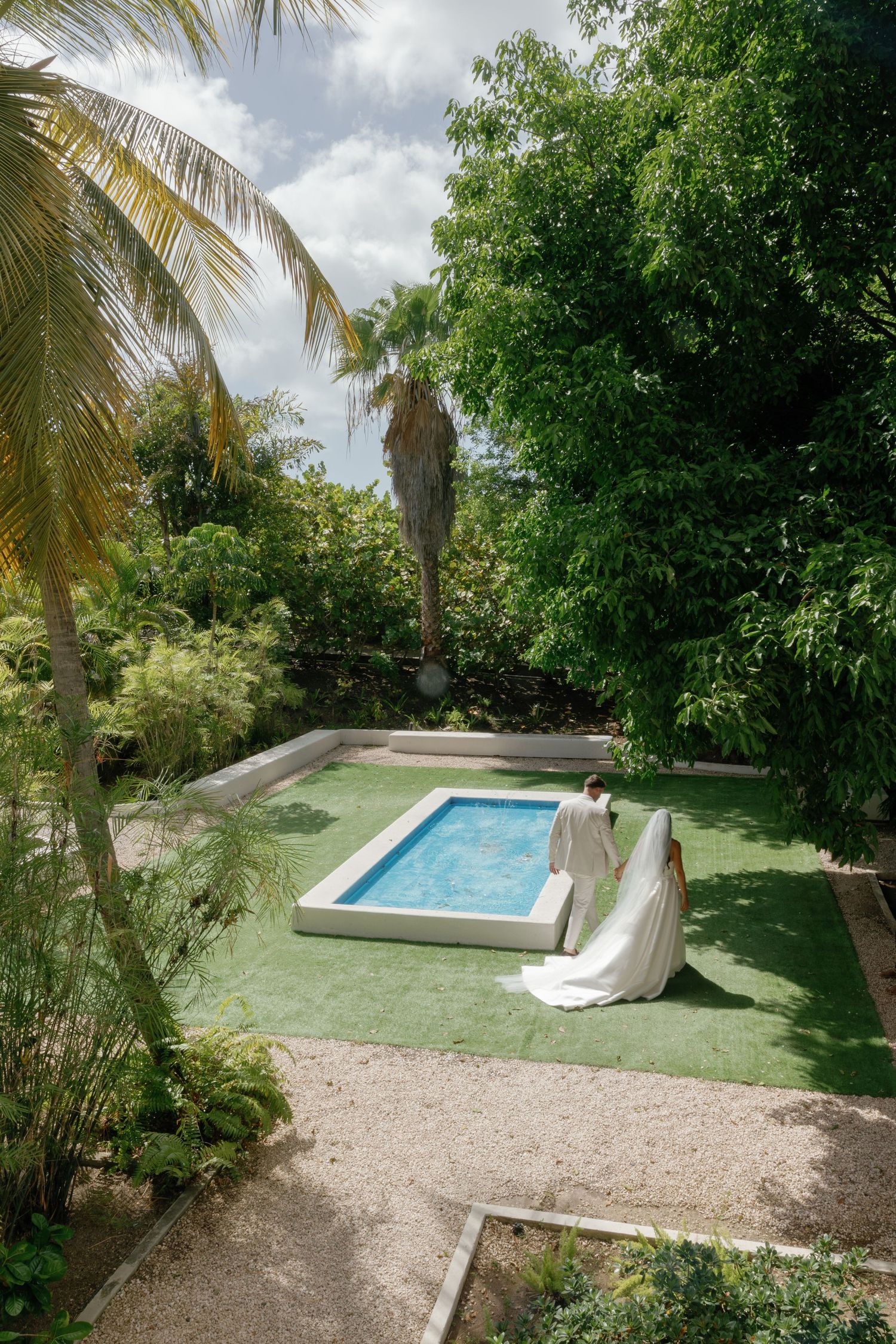 Bride in white gown sitting beside a small rectangular pool surrounded by lush tropical greenery and palm trees.