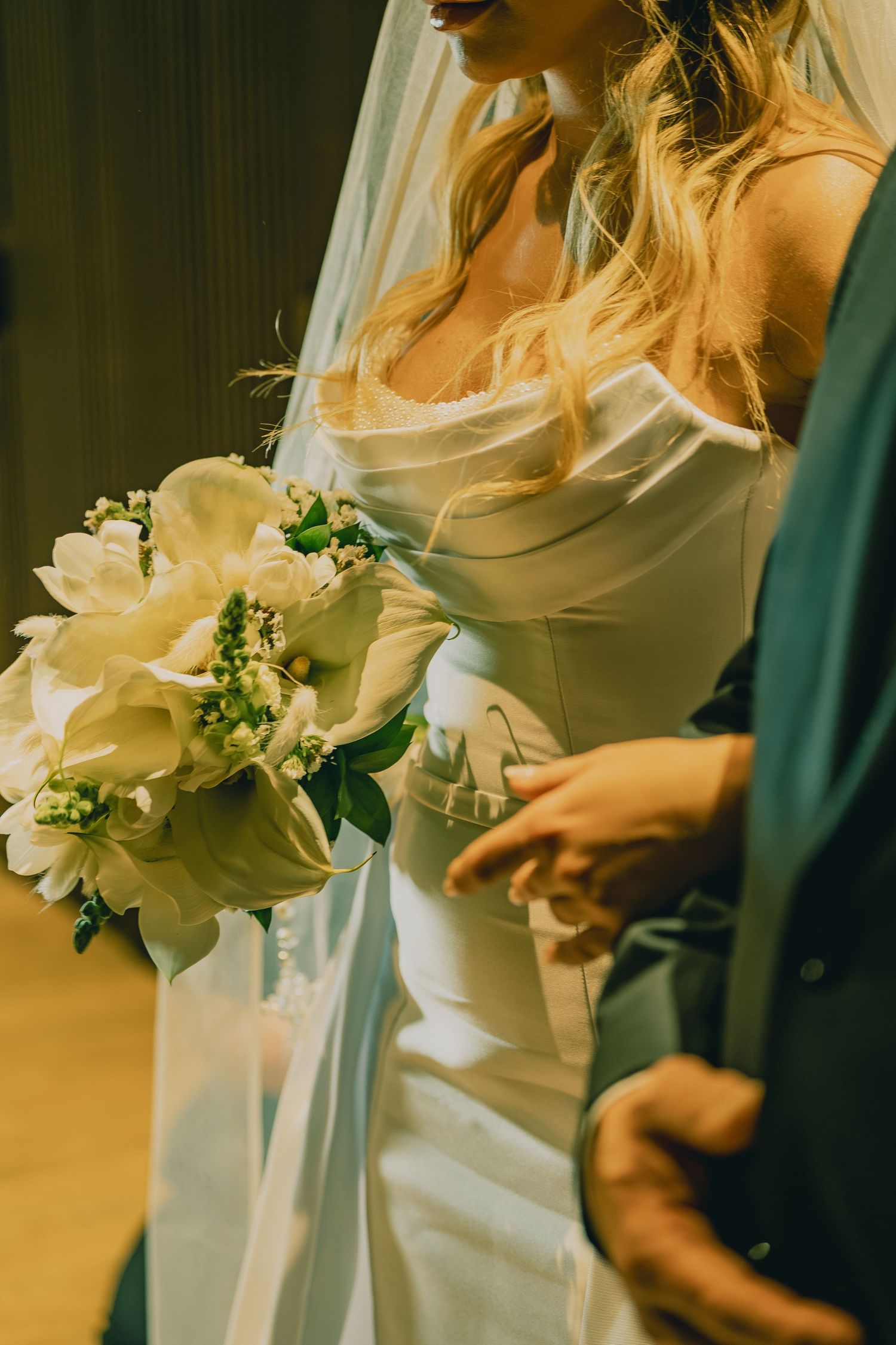 Bride in white satin gown holding elegant white floral bouquet stands beside groom in teal suit at wedding ceremony.