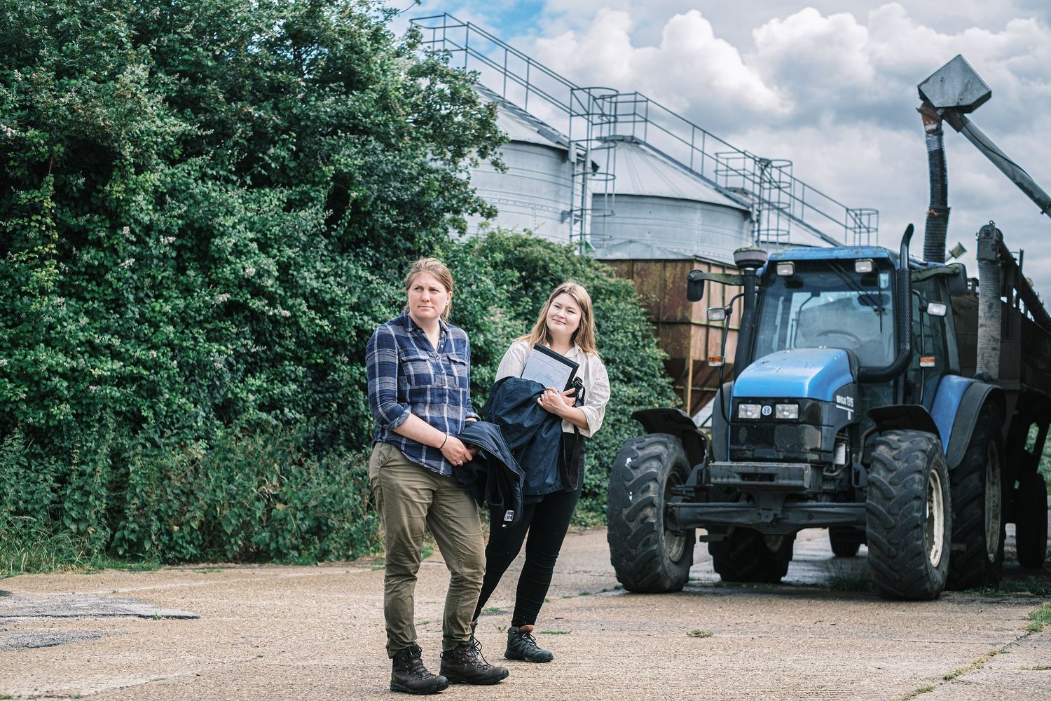 Suffolk Wildlife Trust. Two women stand in front of a blue tractor on a farm with green trees and farm buildings. Photo by John Ferguson