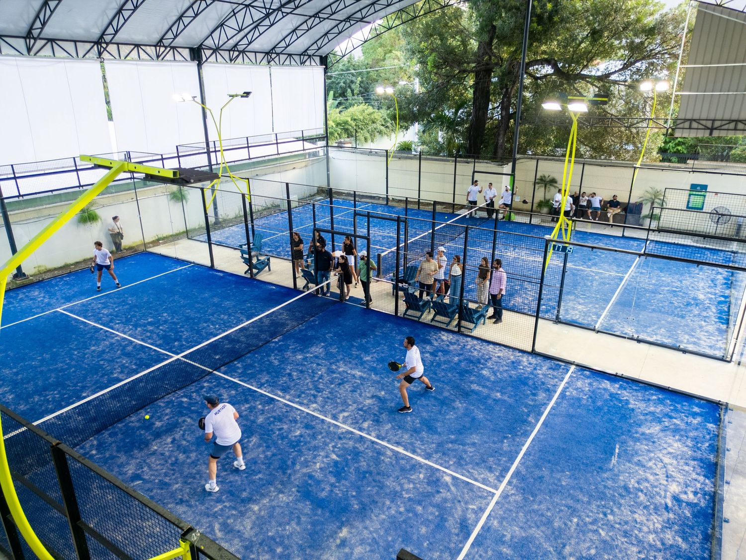 Overhead view of blue padel tennis courts under a covered structure with players and spectators visible.