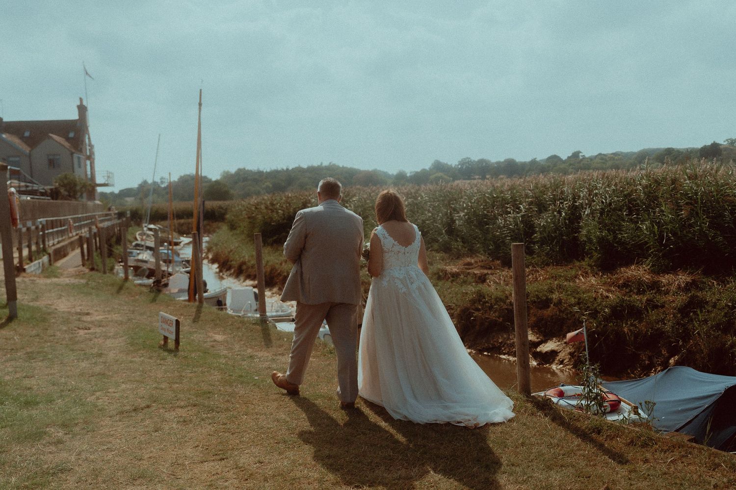 A bride and groom walk hand in hand toward a scenic waterway on their wedding day in a lush countryside setting.