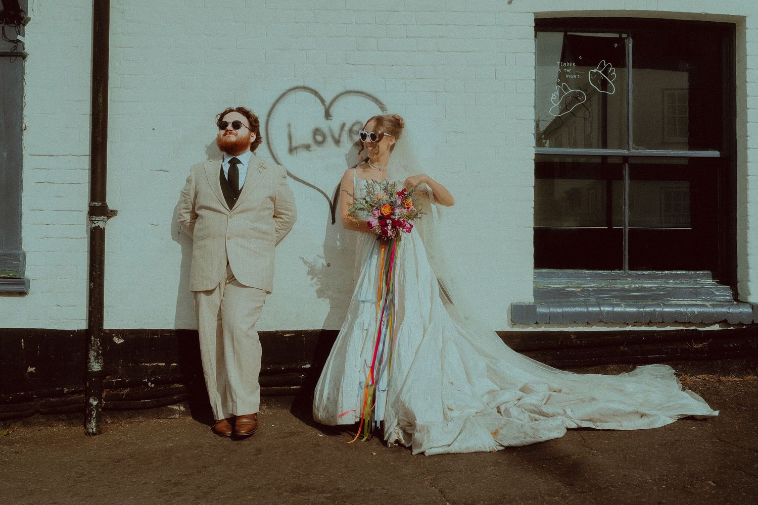 Bride and groom posing against white brick wall with heart and Love graffiti, bride holding colorful bouquet.