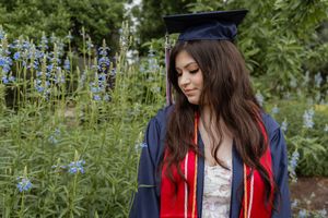 Young woman in graduation cap and gown with red sash poses outdoors among blue wildflowers.