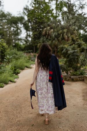 A young woman in a floral dress walks a garden path, carrying her graduation cap and gown.