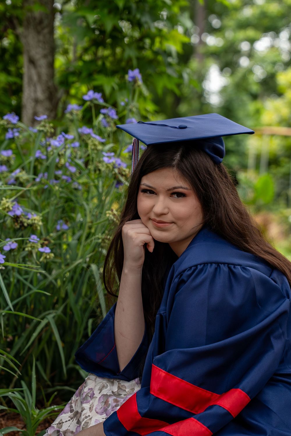 Young woman in navy blue graduation cap and gown poses outdoors among purple flowers and green trees.