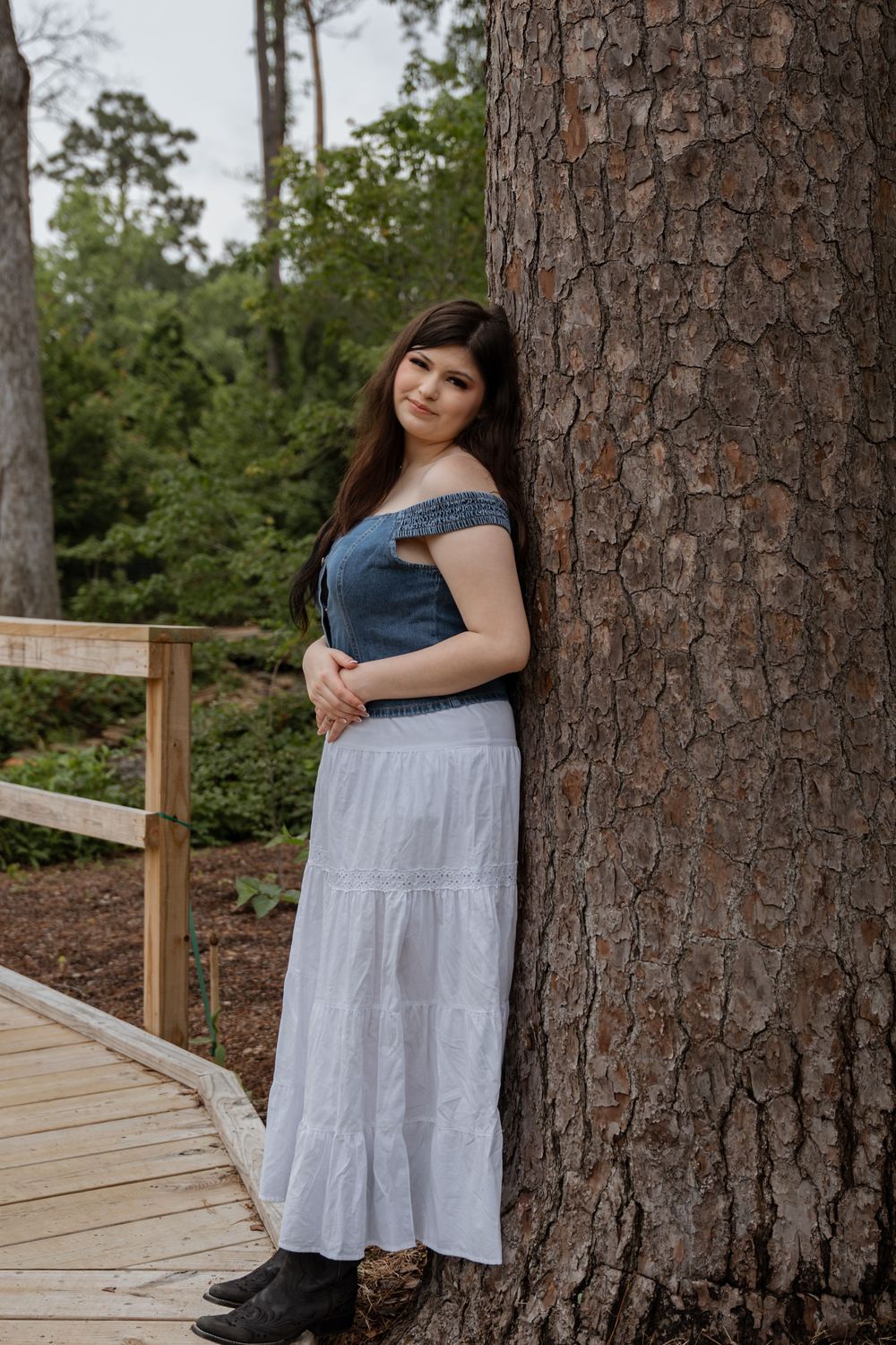 Young woman in blue off-shoulder top and white maxi skirt leaning against a large pine tree on a wooden walkway.