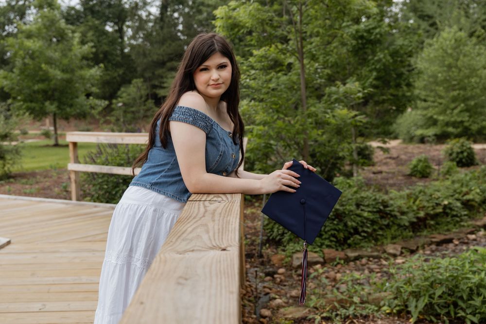 Young woman in denim top and white skirt holding navy graduation cap on wooden bridge in lush garden.