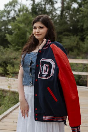 Young woman posing outdoors in a navy and red varsity jacket with letter D patch over a denim vest and white skirt.