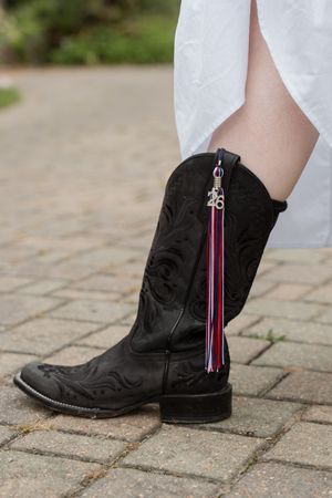 Black cowboy boot with floral embroidery and colorful tassel charm, worn with white dress on brick pavement.