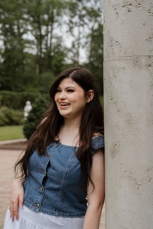 Young woman with long dark hair smiles in denim top and white skirt, leaning against a stone column outdoors.