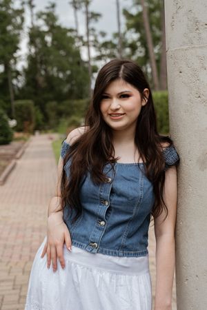 Young woman in denim top and white skirt leaning against a column outdoors, smiling with long dark hair.