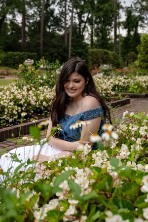 Young woman in blue off-shoulder top sits smiling among white blooming flowers in a lush garden park.