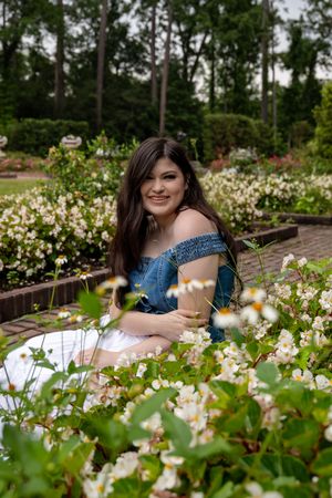 Young woman in off-shoulder denim top smiling among white blooming flowers in a lush garden setting.