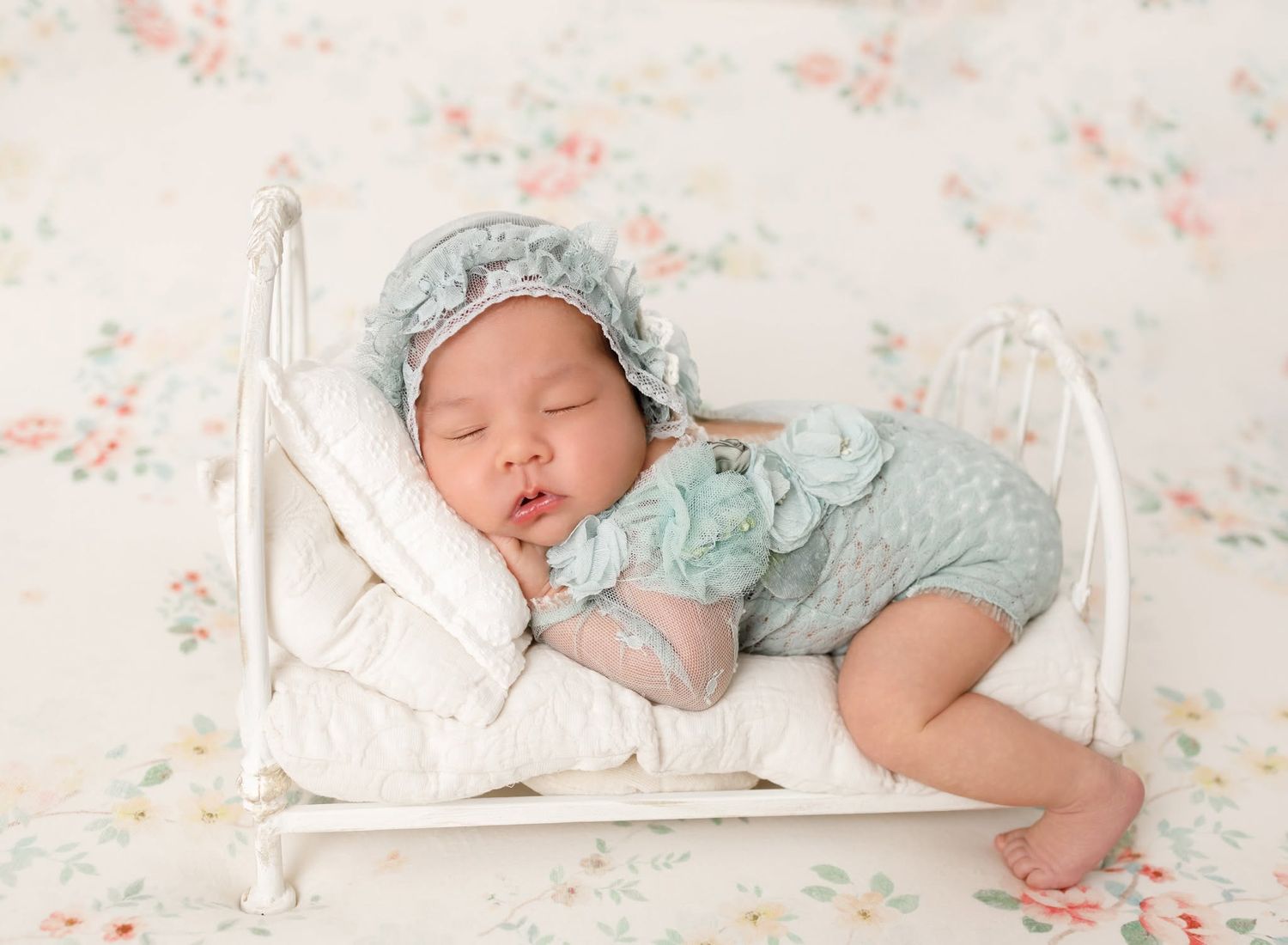 Newborn baby sleeping wrapped in pink swaddle, wearing floral bonnet, holding a small stuffed animal on lace backdrop.