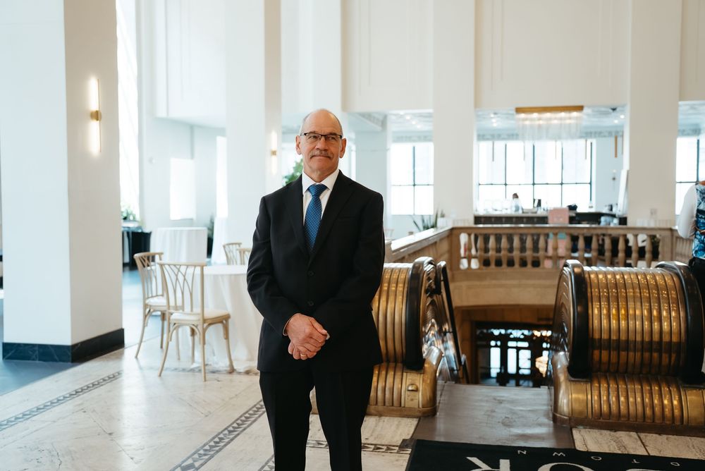 A professional man in a dark suit and blue tie stands confidently in an elegant hotel lobby interior.