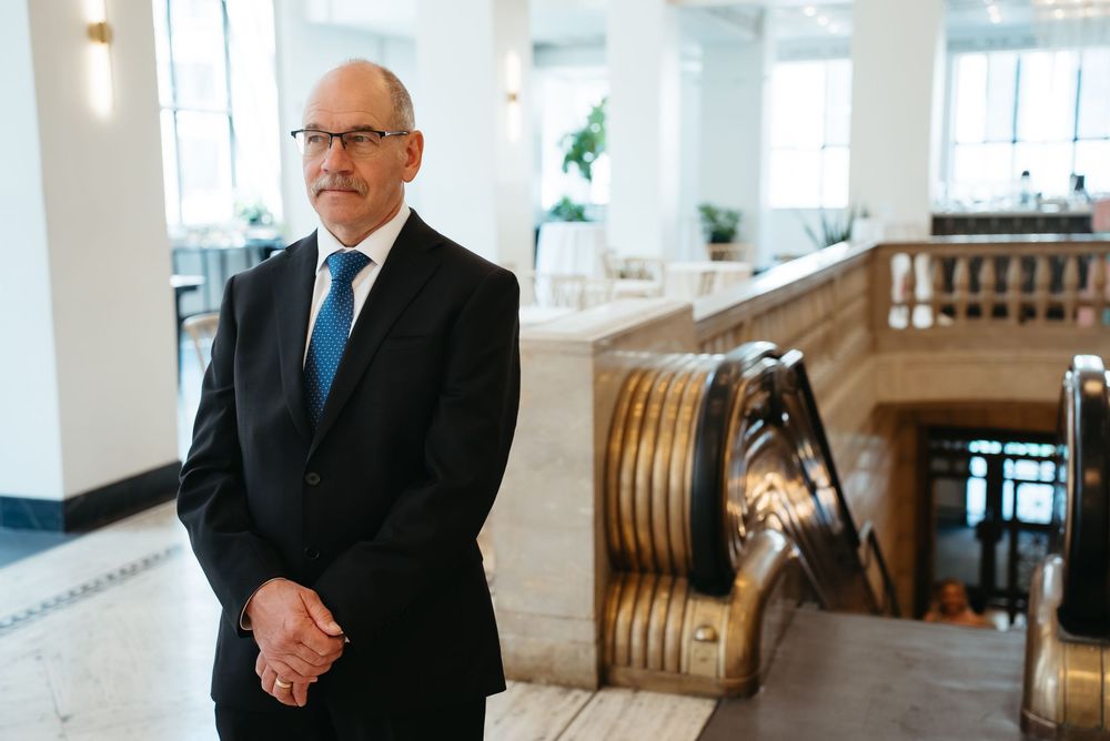 Professional man in dark suit and blue tie standing in elegant building interior with ornate staircase.