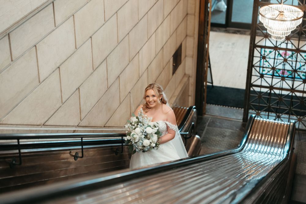 Bride in white ball gown holding bouquet descends ornate staircase in elegant venue.