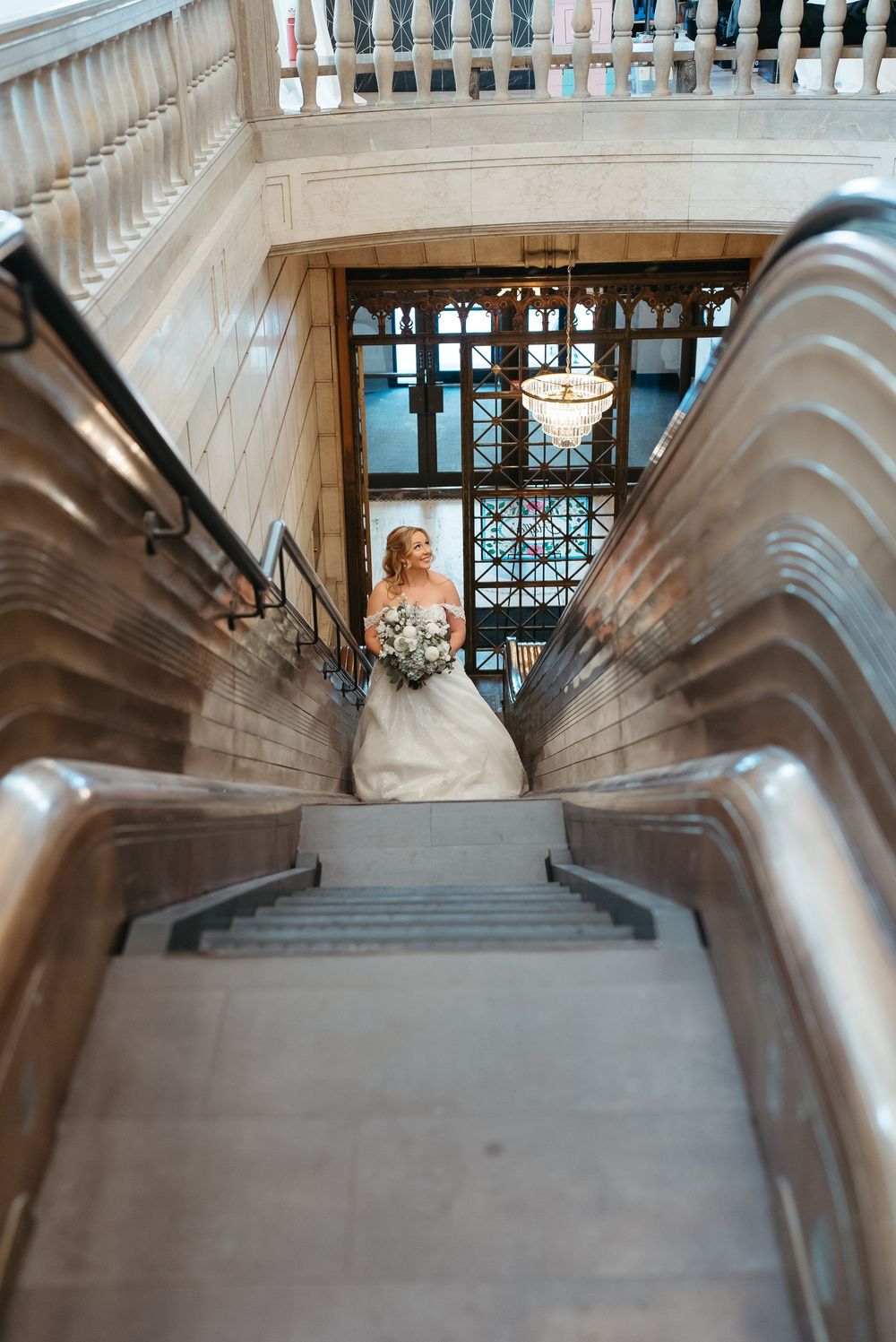 Bride in white gown holding bouquet stands at bottom of grand staircase in elegant venue with chandelier.