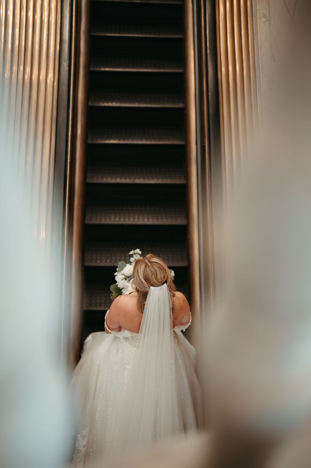 Bride in white gown and floral veil ascending escalator, captured from behind in elegant wedding photography.