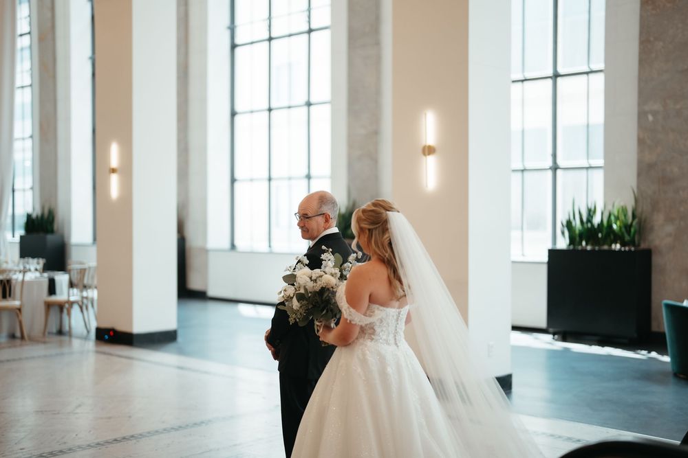 Bride in white gown and veil walks with suited man through elegant venue with tall windows and floral bouquet.