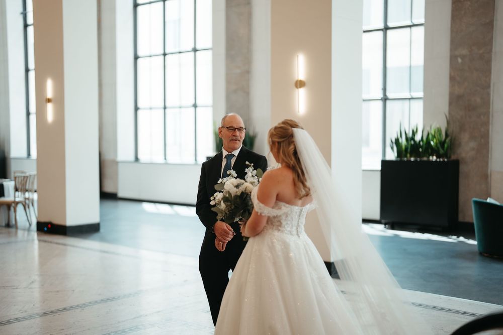 Bride in white gown and veil holding bouquet stands with older man in dark suit in elegant venue.
