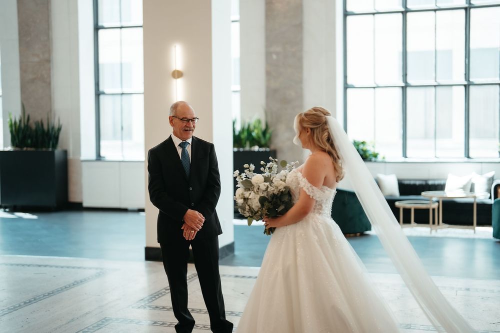 Bride in white lace gown holding bouquet shares emotional first look moment with father in elegant venue.