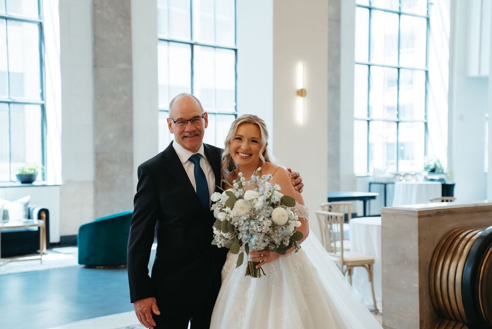 Bride in ball gown holding white bouquet stands with suited man in elegant venue with tall windows.