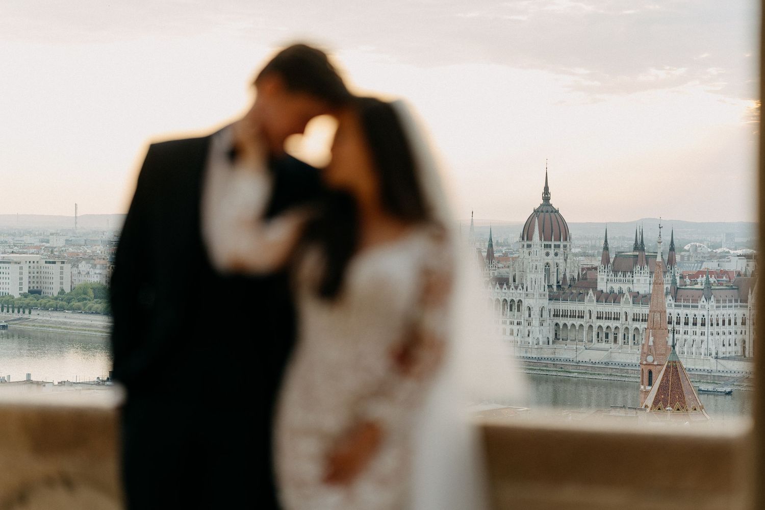 Romantic couple kissing with Budapest Parliament building glowing at sunset in background.