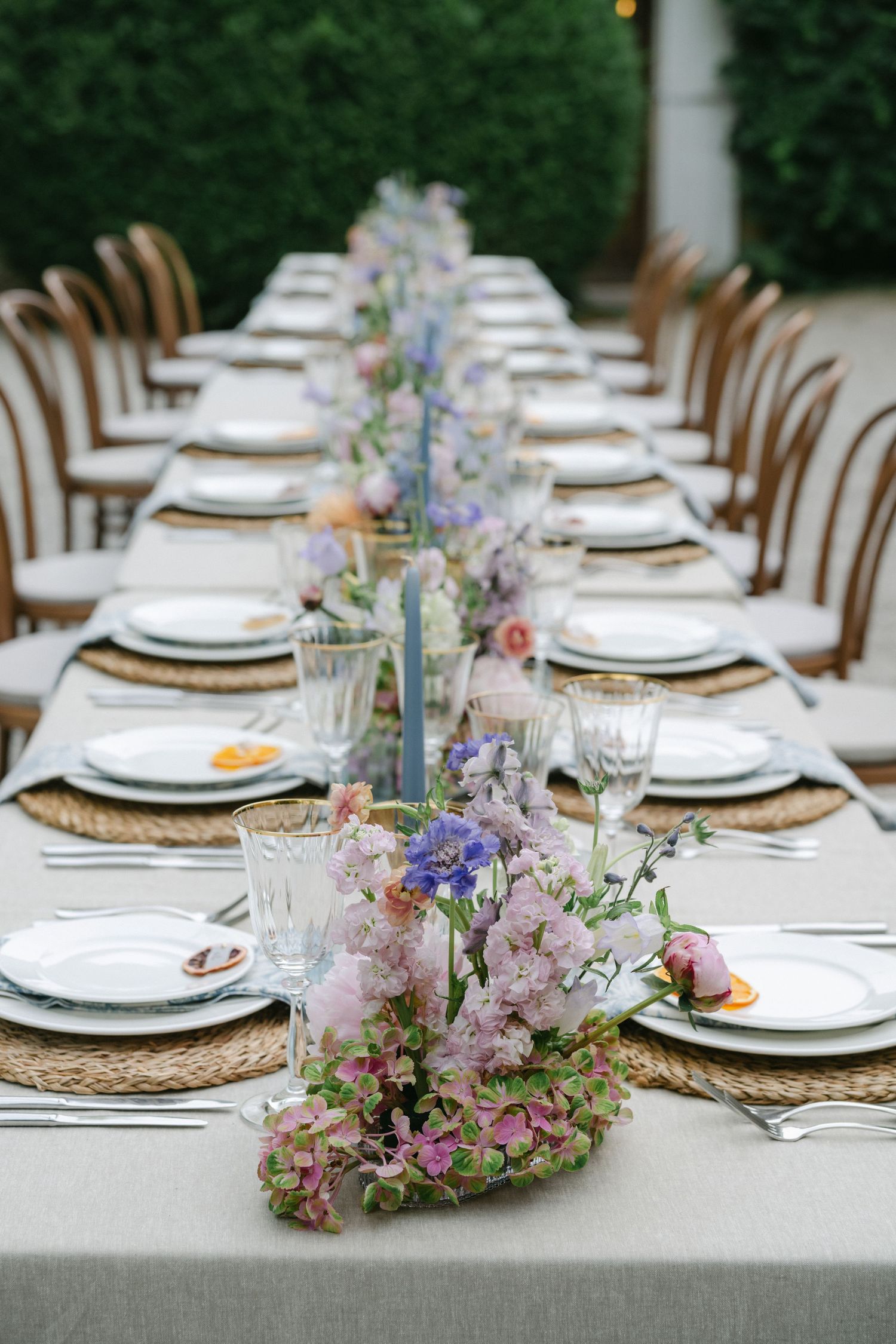 Elegant outdoor wedding reception table with floral centerpieces, wicker chargers, white plates, and glassware on a gray linen tablecloth.