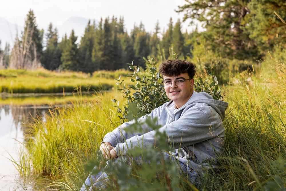 Teen smiling by a mountain pond surrounded by lush green grass and evergreen trees in a scenic wilderness setting.
