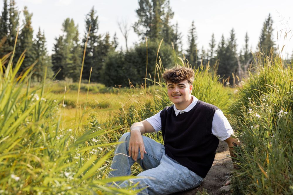 Smiling teen boy sitting in lush green field with tall grass and pine trees in background on sunny day.
