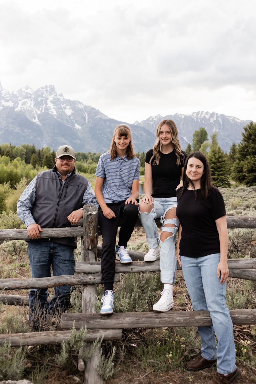 Family of four posing on a rustic wooden fence with snow-capped Grand Teton mountains in the background.
