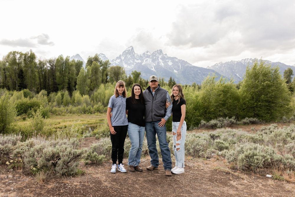 Family of four posing in front of Grand Teton mountains with forest and sagebrush landscape in Wyoming.