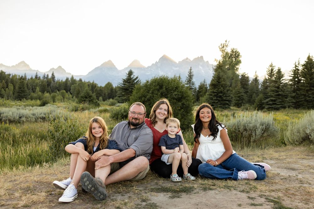 Family of five posing outdoors with Grand Teton mountain range and pine trees in background during golden hour.