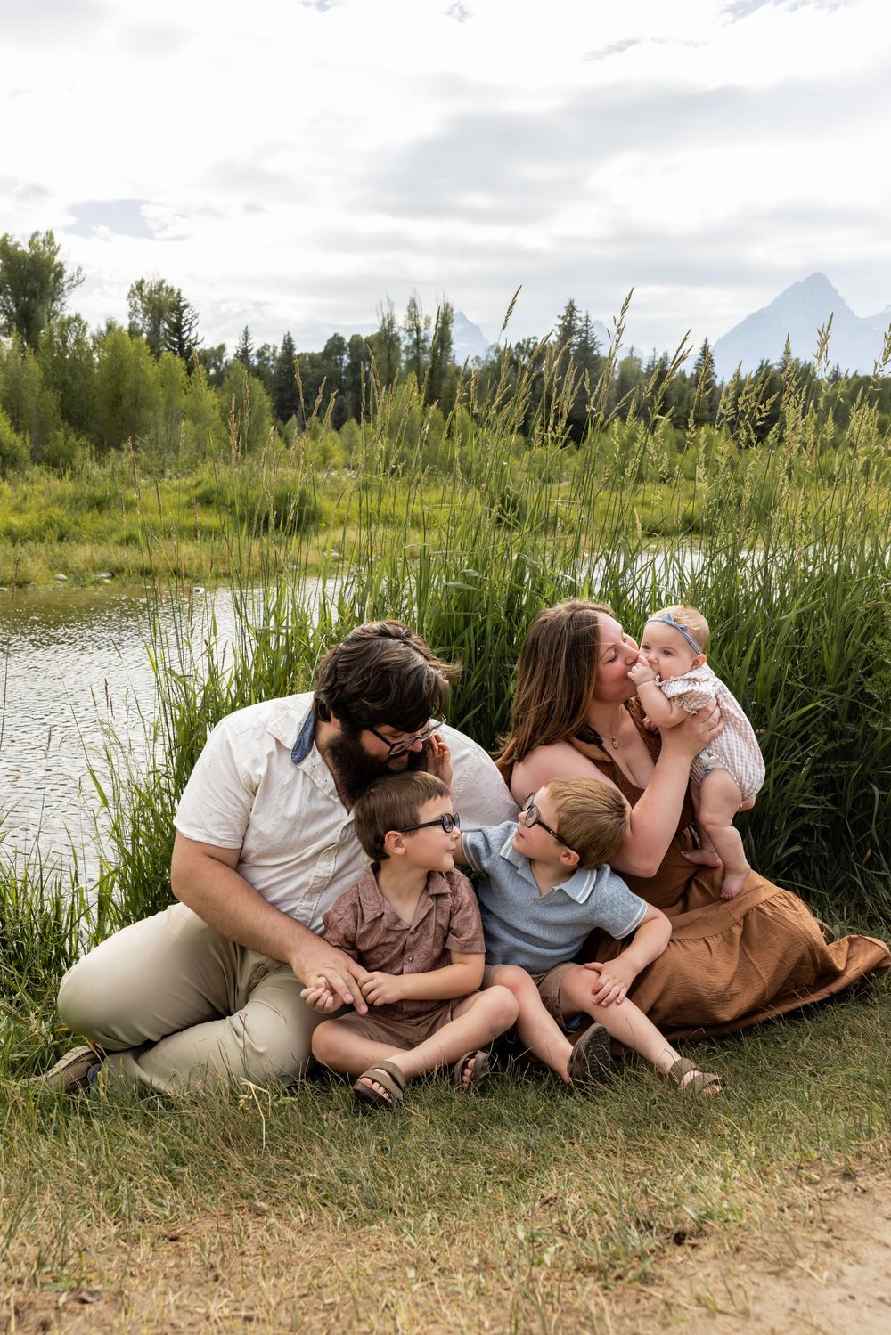 Family of five sitting together by a serene pond with mountain views and lush greenery in the background.